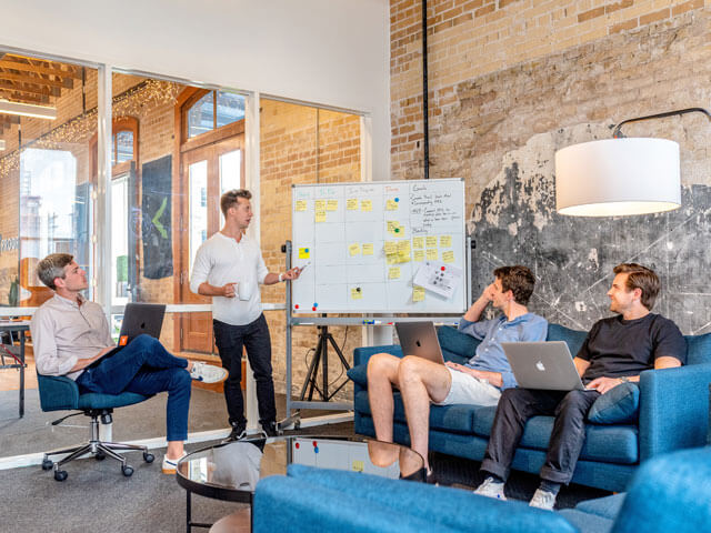 three men sitting while using laptops and watching man beside whiteboard presenting